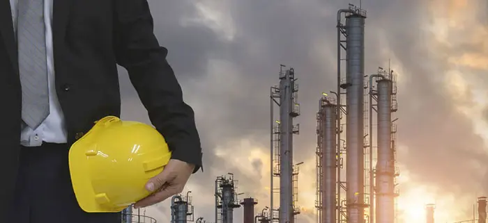 Person in black suit holding yellow hard hat at industrial refinery.